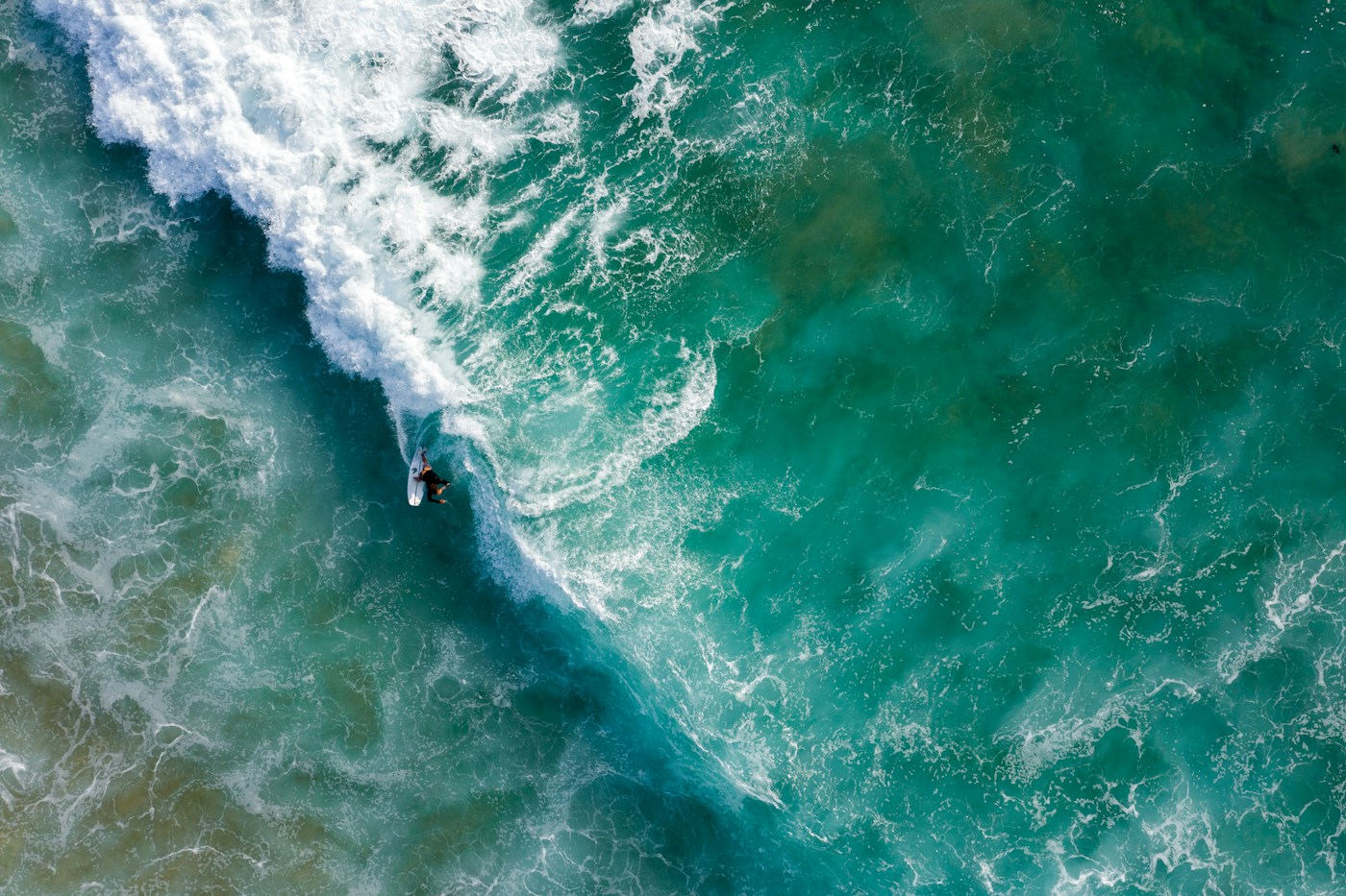 A surfer beside a breaking wave in turquoise water, seen from above
